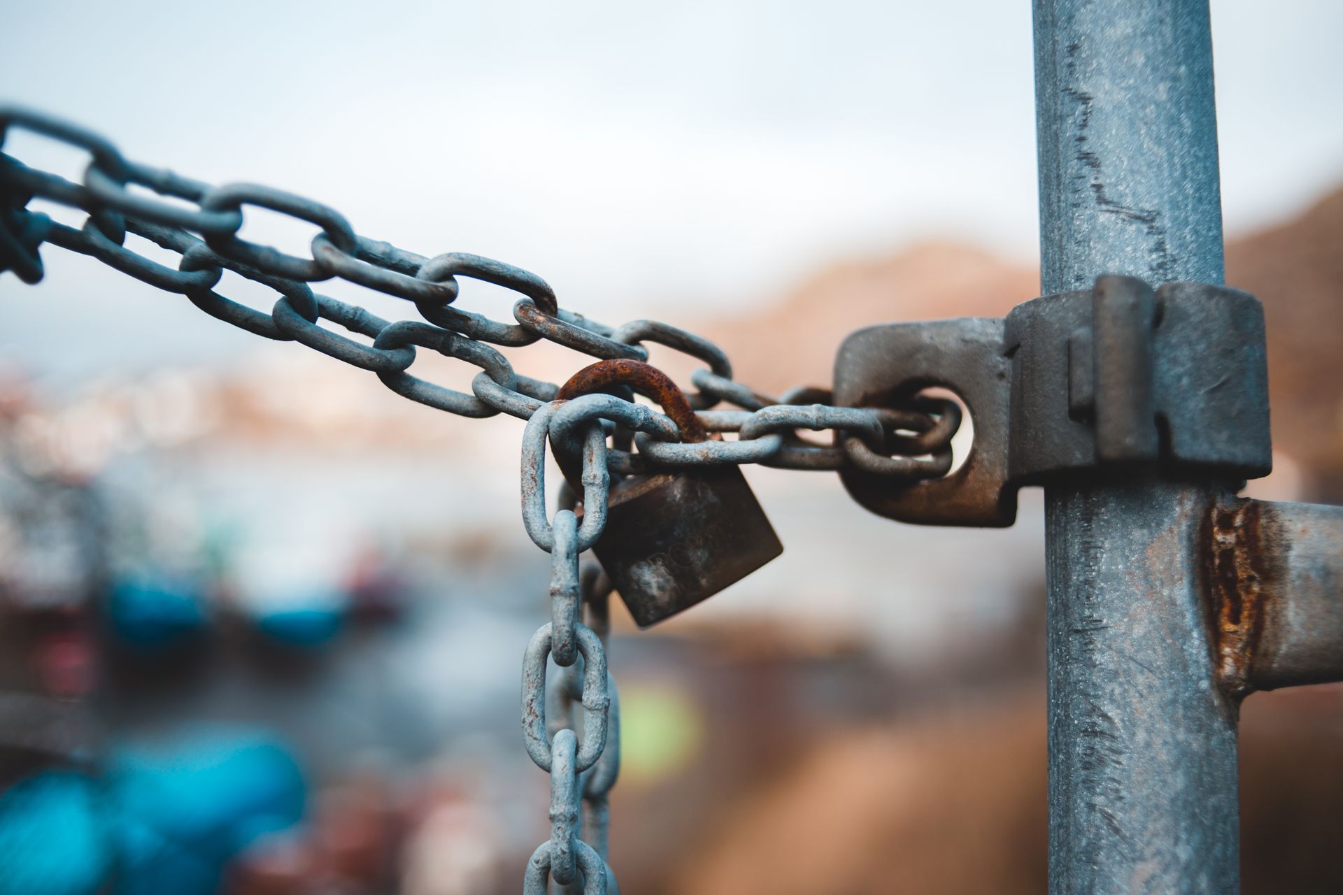 Rusty padlock securing a heavy metal chain to a galvanized fence post with a blurred outdoor background