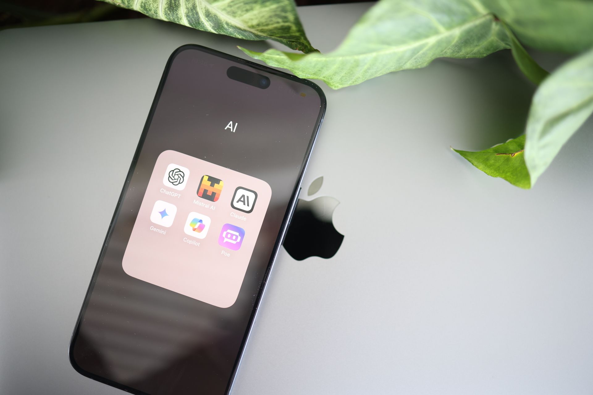 Smartphone resting on a MacBook lid displaying an open AI apps folder with ChatGPT, Claude, and Gemini icons, framed by green plant leaves